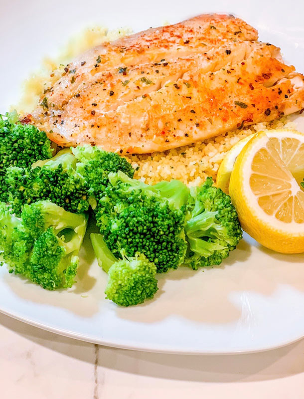 A plate of baked tilapia served a grain and a side of broccoli and a lemon.