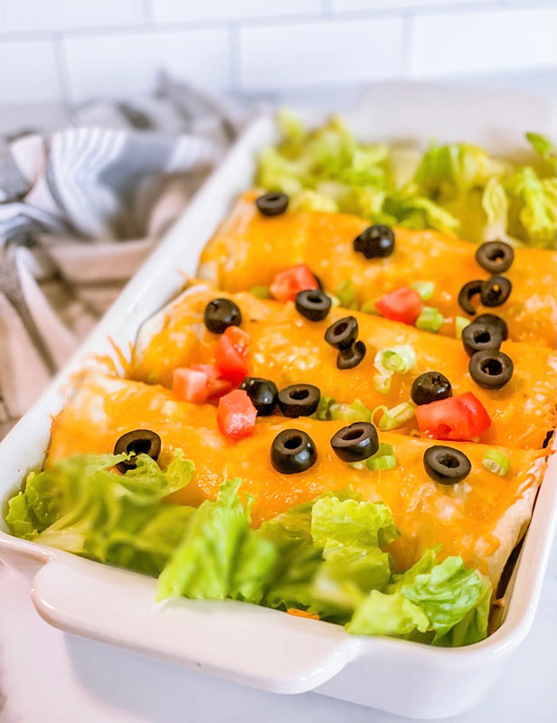 A casserole dish with baked chicken enchiladas with tomatoes and olives on top.