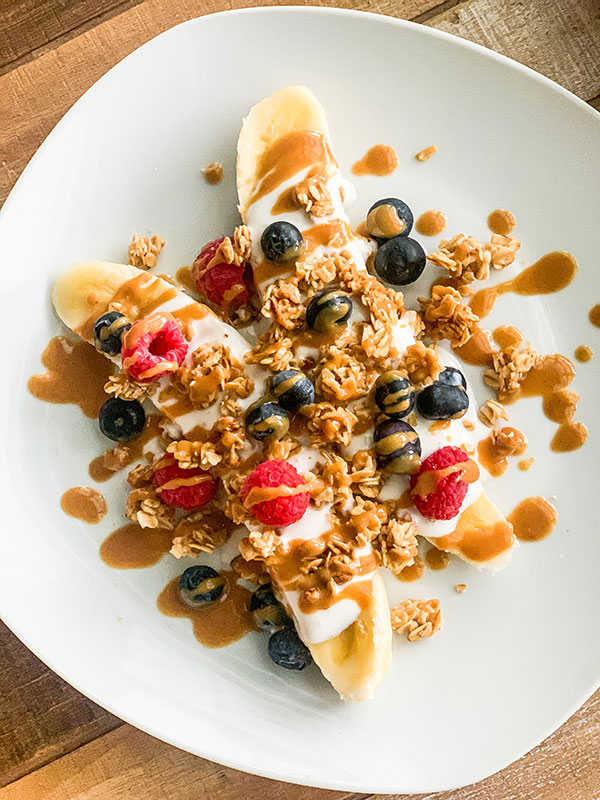 A halved banana on a plate with berries, granola, and drizzles of peanut butter.
